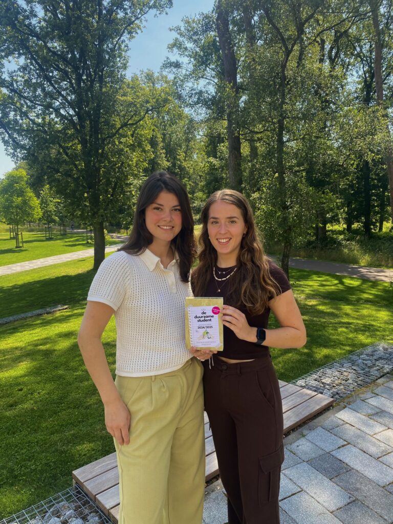 Oud-deelnemers Lily een Judith met hun scheurkalender 'De Duurzame Student'. Lily en Judith staan voor een grasveld met bomen en kijken lachend naar de camera.