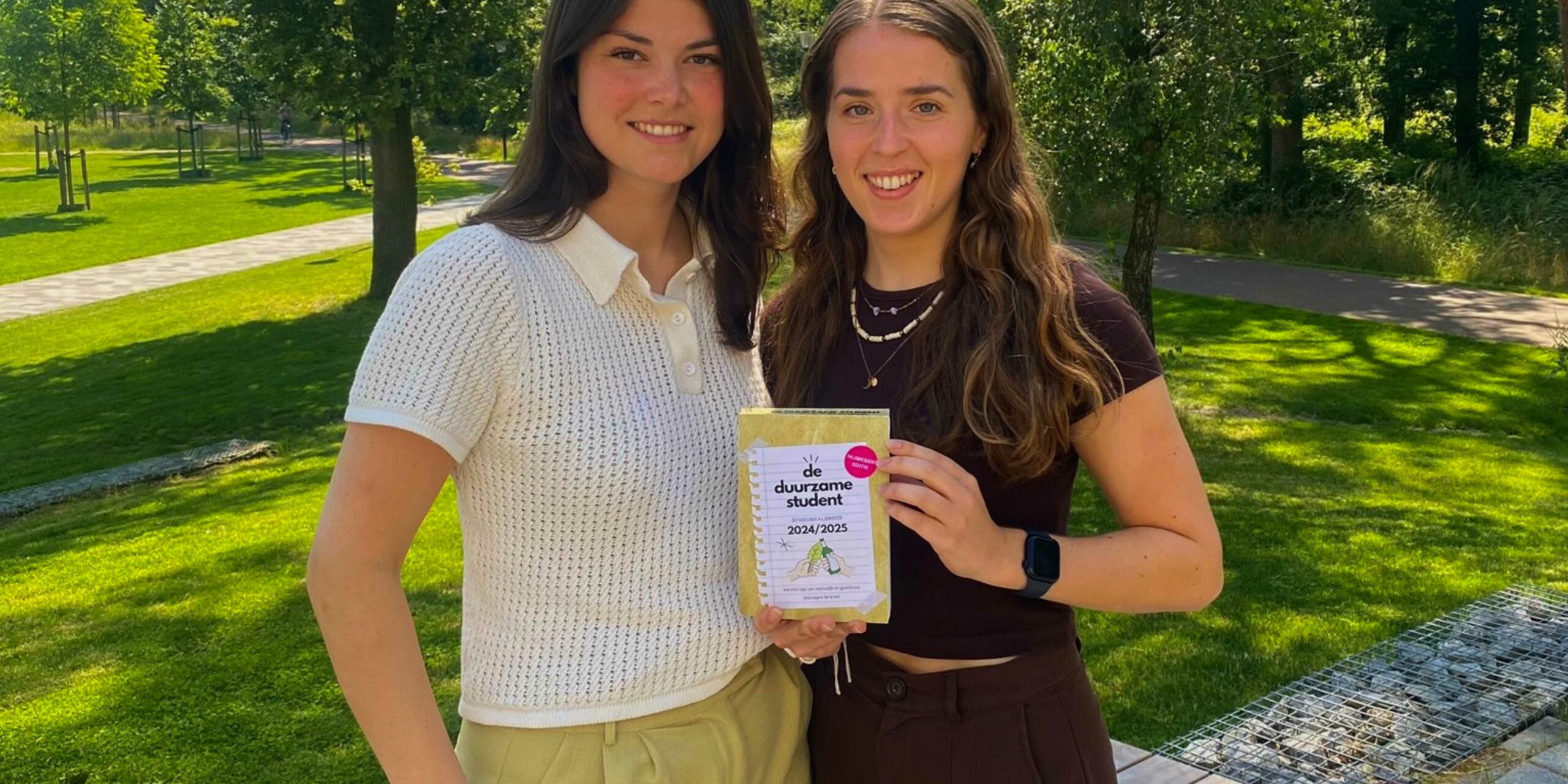 Oud-deelnemers Lily een Judith met hun scheurkalender 'De Duurzame Student'. Lily en Judith staan voor een grasveld met bomen en kijken lachend naar de camera.